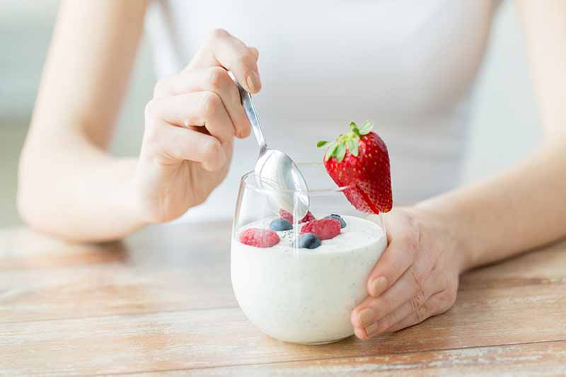 Horizontal image of a woman eating plain yogurt in a glass bowl with fresh fruit at a wooden table.