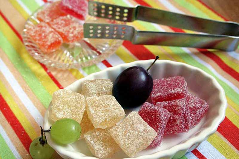 Horizontal image of assorted fruit jelly squares on small plates next to metal tongs.