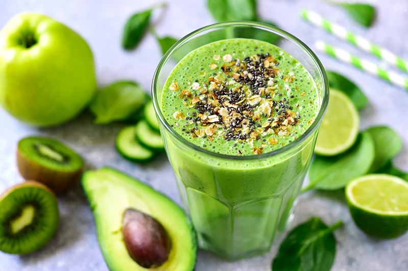 Horizontal image of a creamy green drink next to green fruits and vegetables.