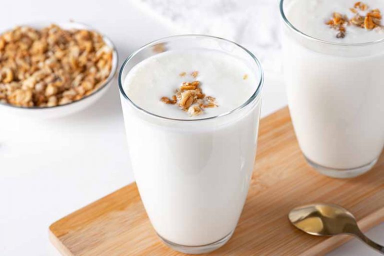 Horizontal image of two glasses filled with a thick white beverage topped with granola on a wooden cutting board.