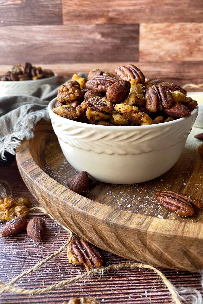 Vertical image of a white bowl filled with seasoned pecans, walnuts, and almonds on a wooden cutting board.