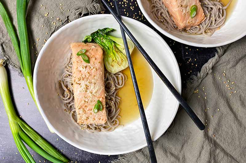 Horizontal image of white bowlfuls of a fish fillet on top of noodles and broth with chopsticks next to scallions.