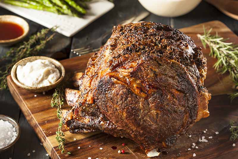 Horizontal image of a large piece of meat with a browned exterior on a wooden board next to horseradish sauce.