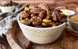 Horizontal image of a white bowl filled with seasoned pecans, walnuts, and almonds on a wooden cutting board.