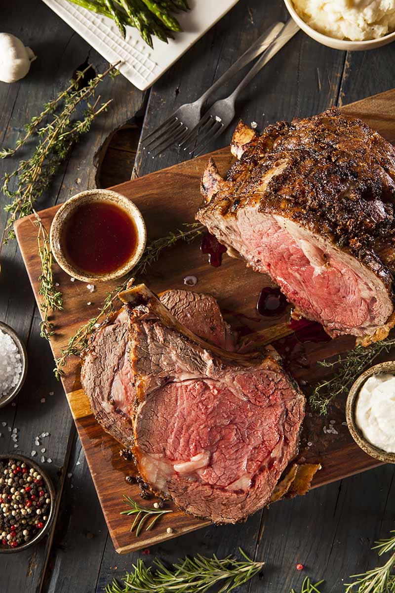 Vertical top-down image of a large cut of cooked rare beef on a wooden board next to bowls of sauces, herbs, and vegetables.