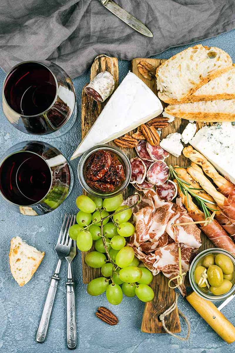 Vertical image of a meat, cheese, nut, and fruit plate next to glasses of wine.