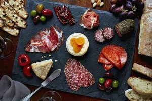 Horizontal image of a slate platter with assorted sliced charcuterie and other accompaniments.