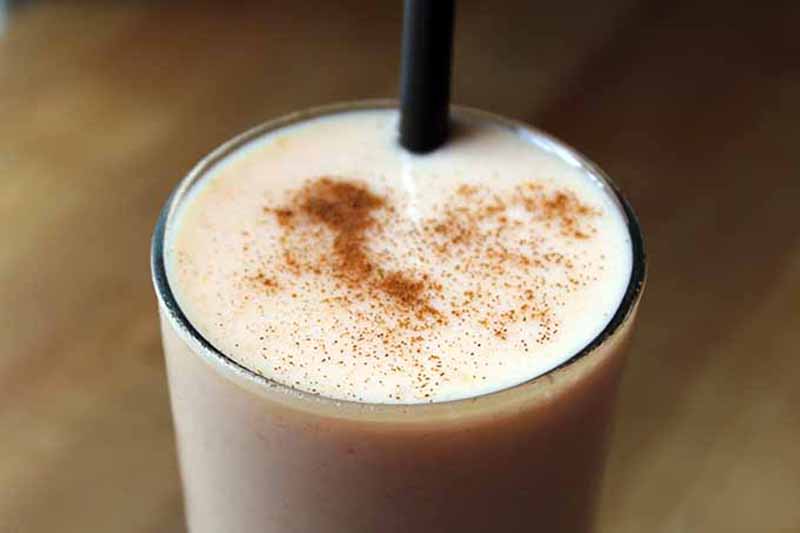 Horizontal close-up image of a glass filled with a light orange-colored smoothie topped with cinnamon with a black straw inserted.