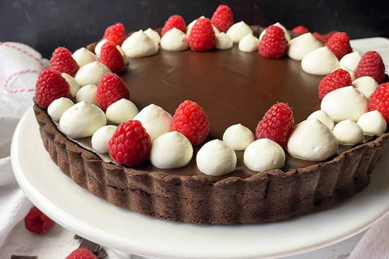 Horizontal image of a dessert with a cocoa crust garnished with whipped cream dollops and raspberries on a white cake stand.