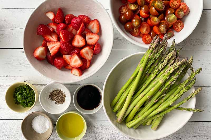 Horizontal image of sliced strawberries and tomatoes next to whole green vegetable spears and small bowls of seasonings.