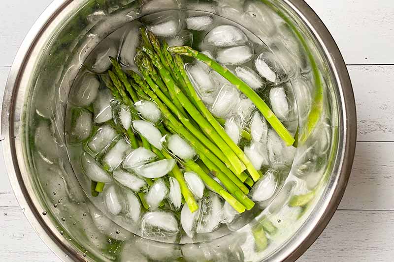 Horizontal image of green veggie spears in a large bowl filled with ice water.