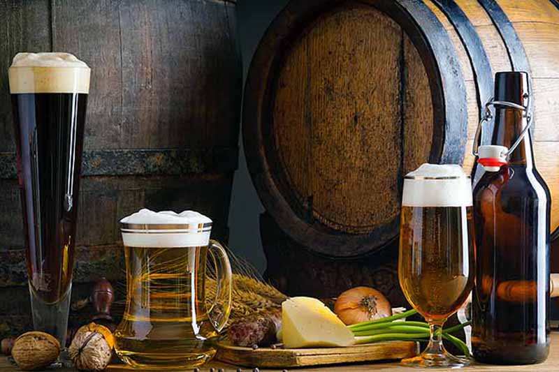 Horizontal image of assorted glasses of beer next to food and a barrel.