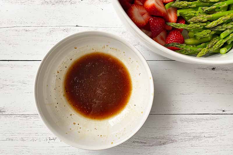 Horizontal image of a small bowl filled with a dark brown dressing next to a larger bowl with fruit and vegetables.