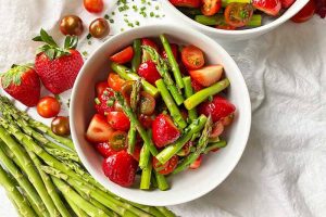 Horizontal image of two white bowls filled with a mix of green vegetables, strawberries, and tomatoes.
