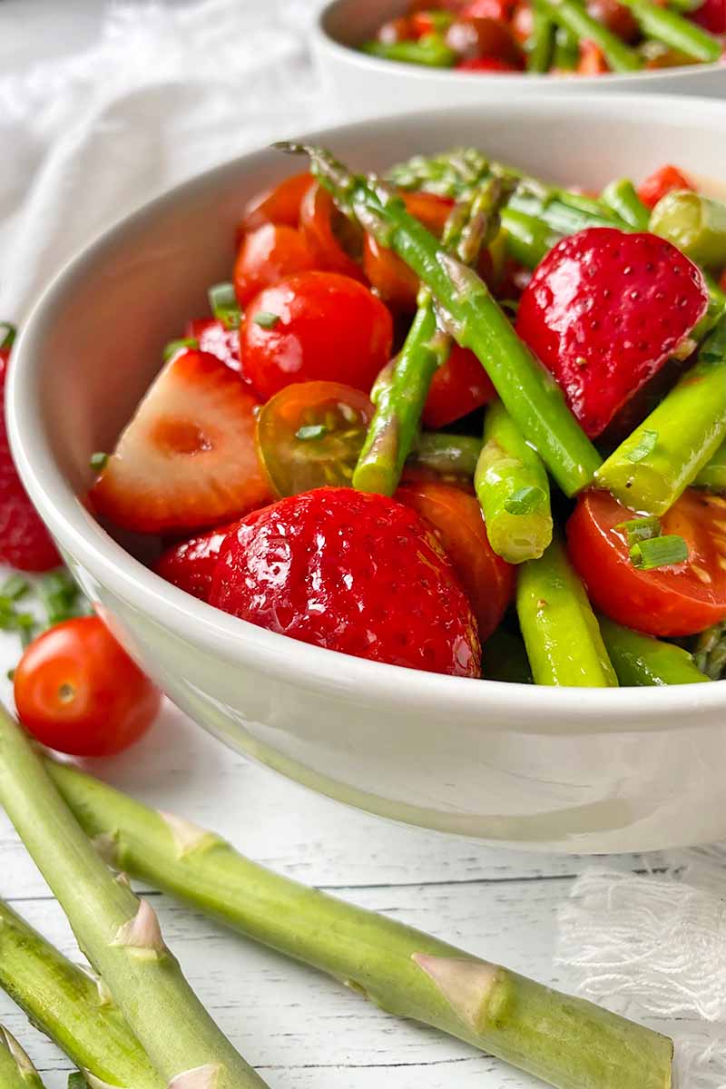 Vertical close-up image of a mix of bright green vegetable spears with strawberries and tomatoes in a white bowl.