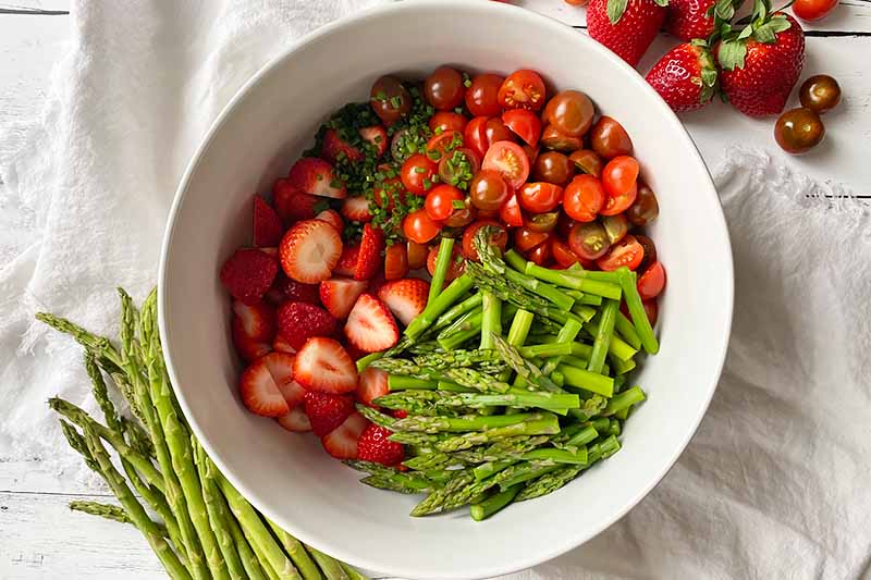 Horizontal top-down image of a large white bowl with sliced strawberries, tomatoes, and green stalks.