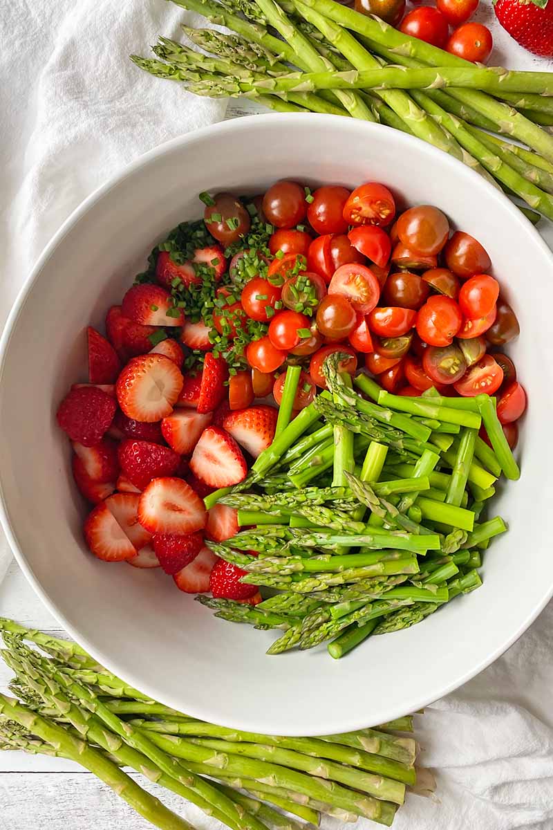 Vertical top-down image of a large white bowl with sliced strawberries, tomatoes, and green stalks.