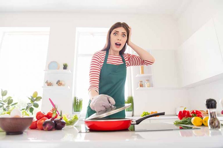 Horizontal image of a shocked woman opening the lid to a pot in the kitchen.