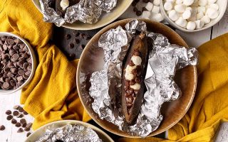 Horizontal image of a plate with a stuffed roasted whole fruit in aluminum foil next to yellow towels and chocolate chips.