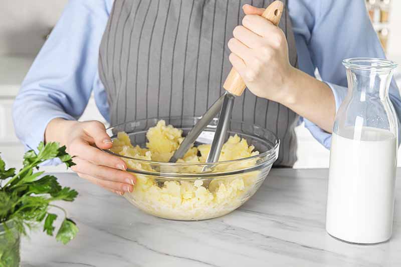 Horizontal image of mashing potatoes with a masher in a clear glass bowl.
