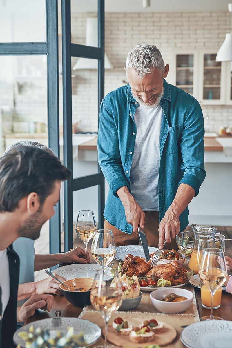Vertical image of an older man carving a roast chicken during a dinner party.