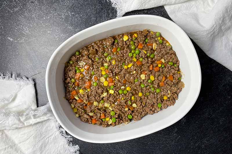Horizontal image of a ground lamb and mixed vegetable mixture spread on the bottom of a ceramic white baking dish.