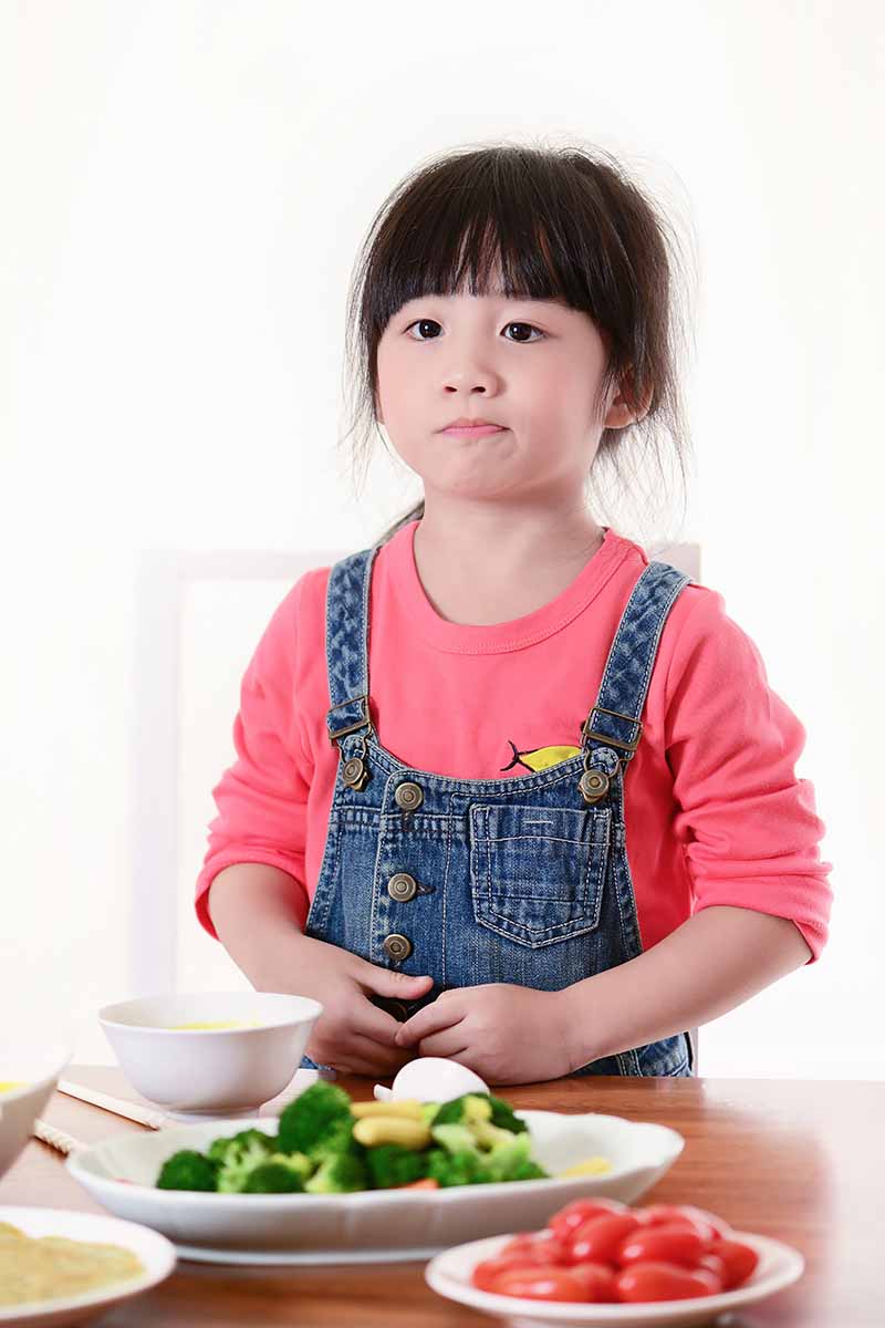 Vertical image of a young girl refusing to eat raw vegetables on white plates at a table.