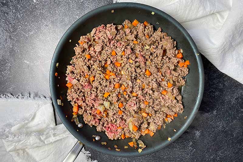 Horizontal image of cooking ground beef with diced carrots and onions in a skillet.