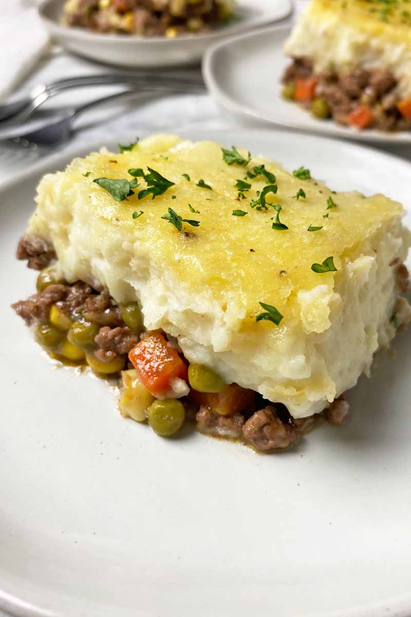 Vertical close-up image of a large serving of a layered shepherd's pie recipe with ground lamb and assorted vegetables on a white plate.