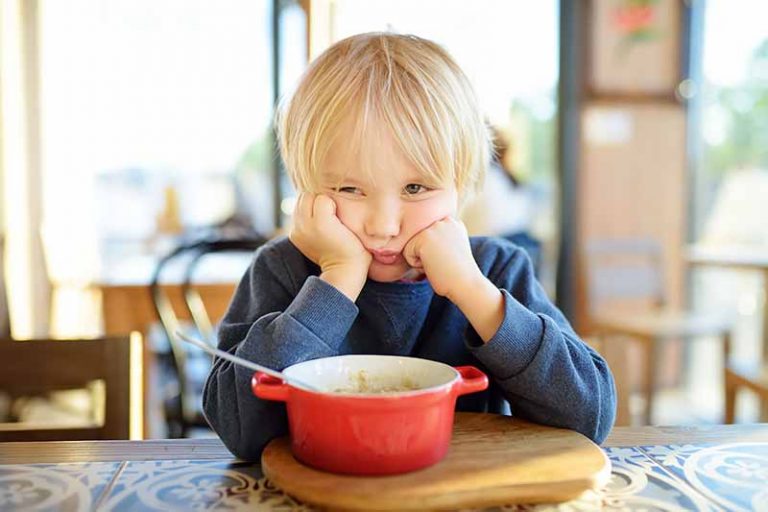 Horizontal image of a cranky child sitting at the table with a red bowl.