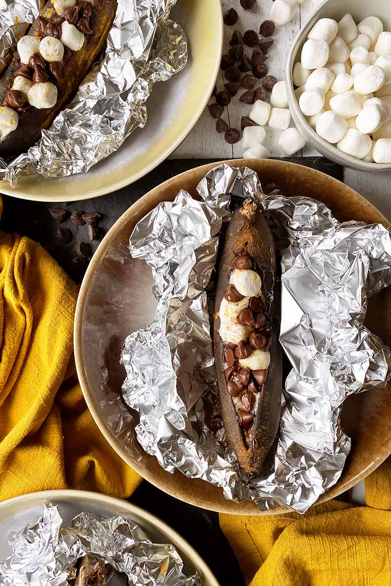 Vertical image of a plate with a stuffed roasted whole fruit in aluminum foil next to yellow towels and mini marshmallows.