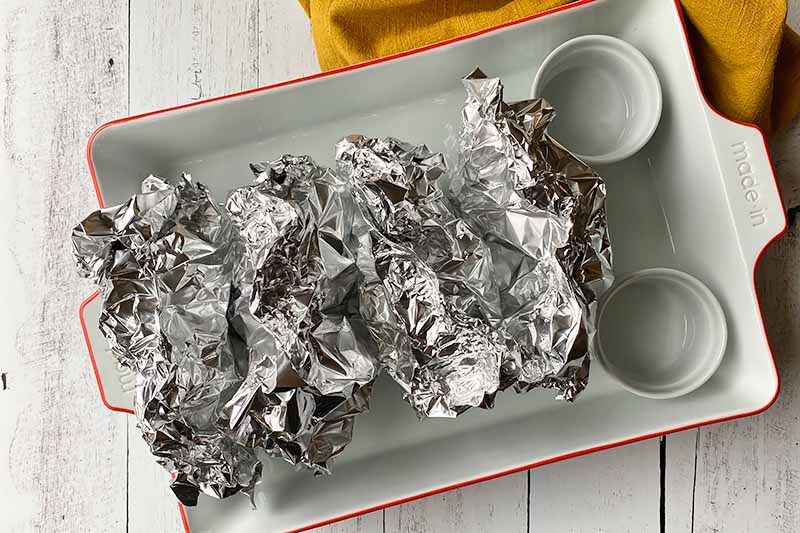 Horizontal image of four packages wrapped in aluminum foil in a ceramic baking dish.