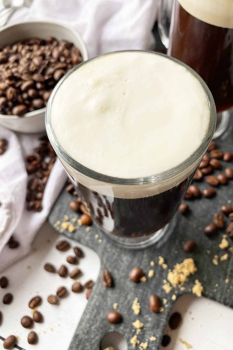 Vertical image of a glass filled with a deep brown beverage with a whipped topping surrounded by java beans.
