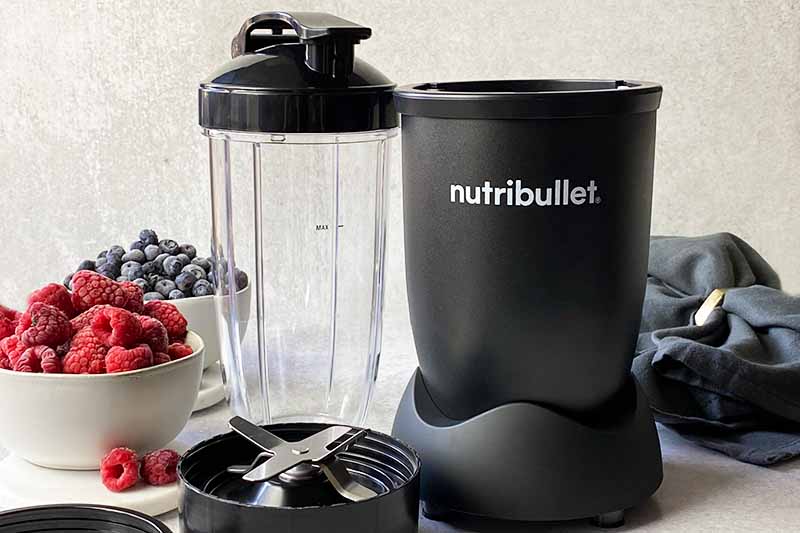 Horizontal image of the Nutribullet blender and its accessories next to bowls of fresh berries.