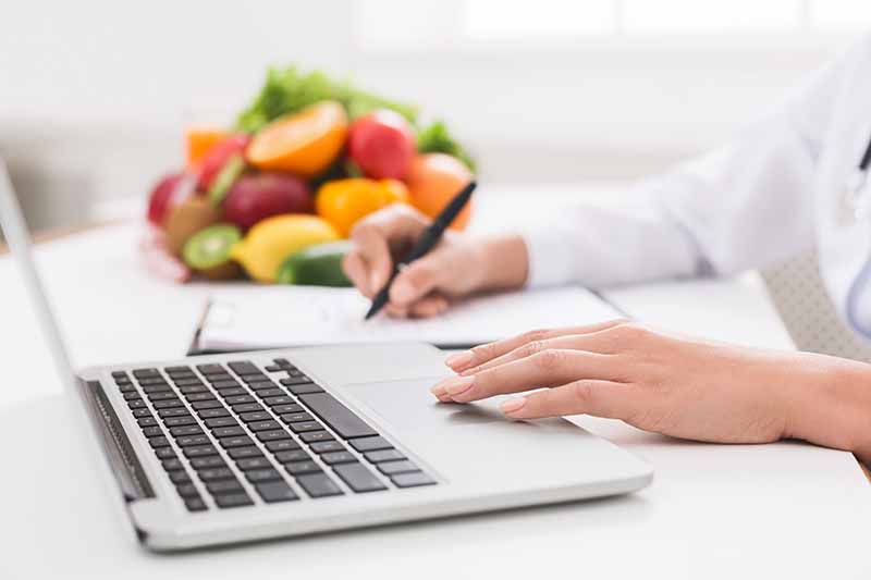 Horizontal image of writing on a pad of paper and typing on a computer next to whole produce.