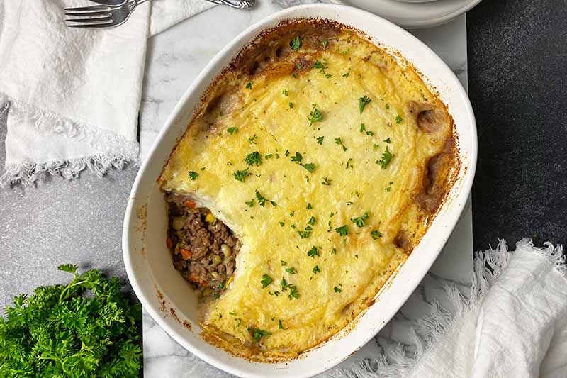 Horizontal top-down image of a whole shepherd's pie with a browned crust and a ground meat filling next to parsley.
