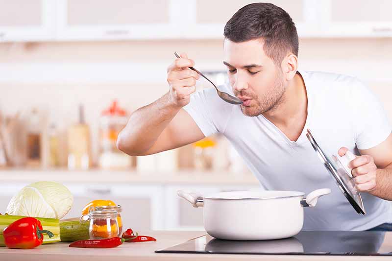 Horizontal image of a man tasting food in a pot over a stovetop.