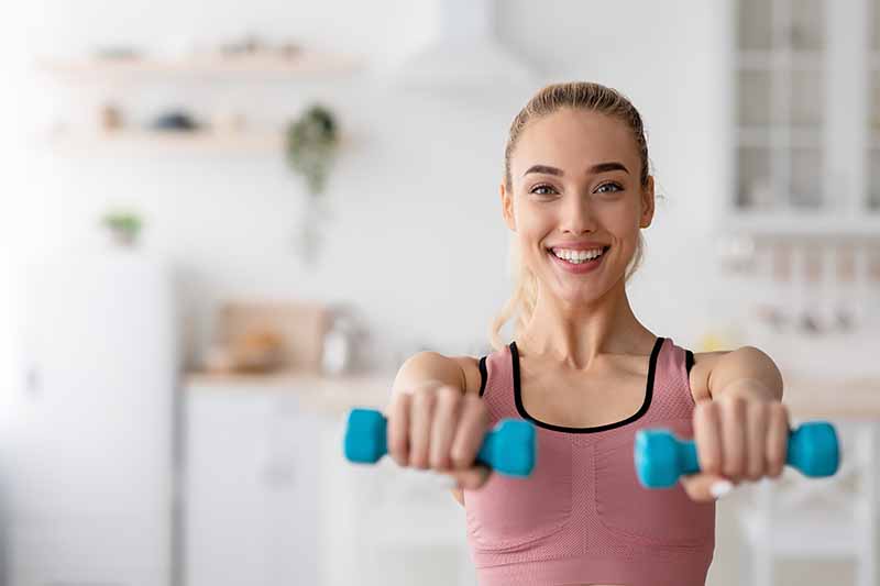 Horizontal image of a woman lifting small blue weights at home.