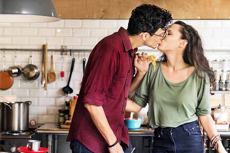 Horizontal image of a man and women kissing while eating spaghetti.