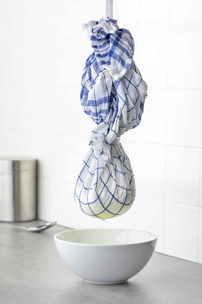 Vertical image of straining food in a cloth over a white bowl on a metal counter.