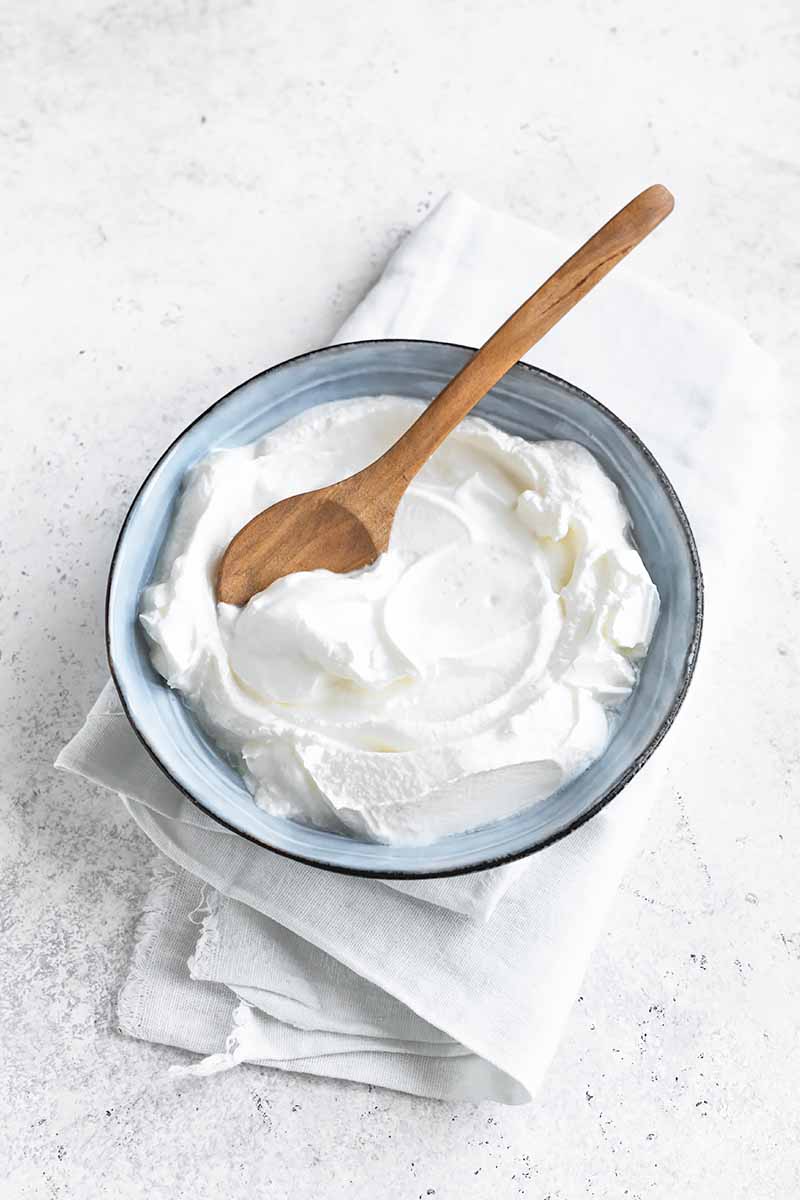 Vertical image of a thick and spreadable white cream in a blue bowl next to a wooden spoon.