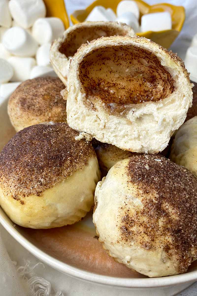 Vertical image of rolls coated in cinnamon sugar in a bowl, with one sliced in half to reveal a hollow inside.