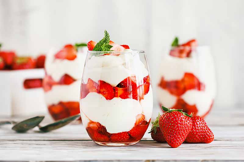 Horizontal image of multiple strawberry parfaits in glasses on a wooden cutting board on a white table.