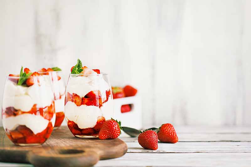Horizontal image of multiple strawberry parfaits in glasses on a wooden cutting board on a white table.