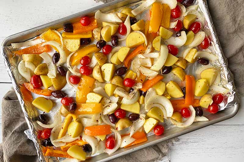 Horizontal image of a mix of seasoned raw prepped vegetables, whole tomatoes, and whole olives on a baking sheet.