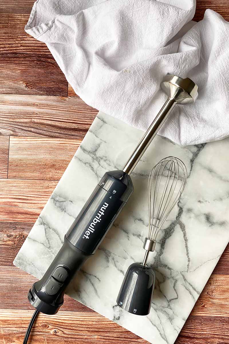 Horizontal top-down image of a narrow handheld kitchen appliance next to a whisk accessory on a marble slate and white towel.
