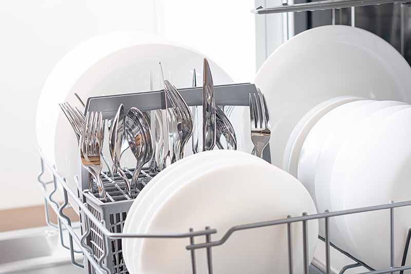 Horizontal image of plates and silverware organized on a rack in a kitchen cleaning appliance.