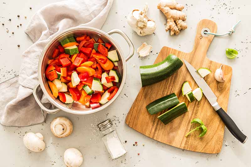 Horizontal image of prepped zucchini on a wooden cutting board next to a knife and a pot of vegetables.