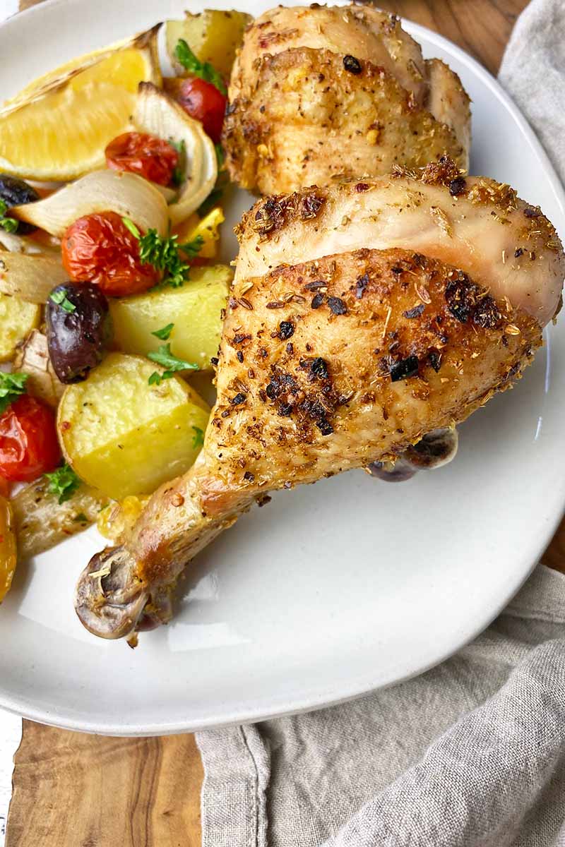Vertical image of two crispy drumsticks next to quartered potatoes, tomatoes, and olives on a white plate.