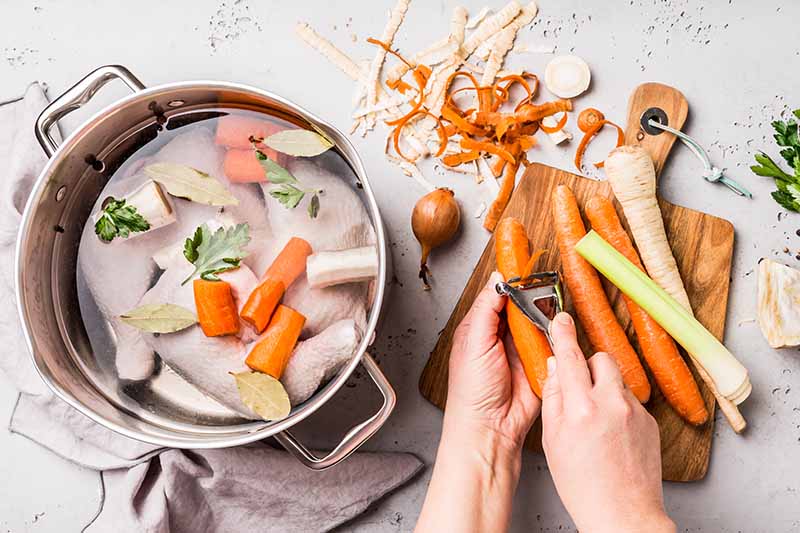 Horizontal image of hands peeling carrots next to a pot with soup and vegetables in it.